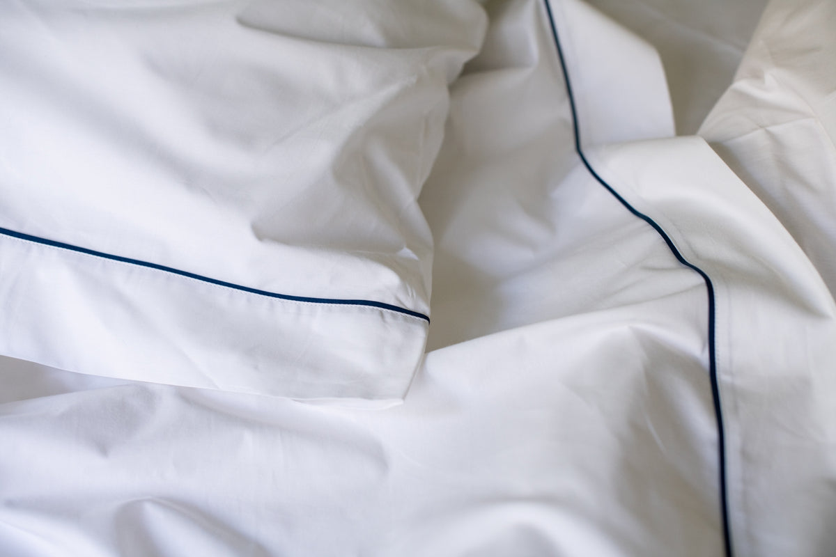 Close-up of white bed sheet with navy piping detail, showing smooth fabric